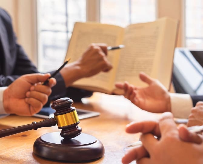 A group of people discuss over an open book at a table. One person holds a pen; another's gavel lies on the table.