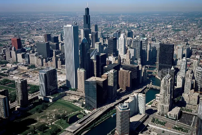 Aerial view of a dense cityscape featuring tall skyscrapers, intersecting bridges, and a meandering river, with sprawling urban areas in the background under a clear sky.