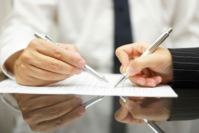 Two people wearing formal attire are seated at a reflective table, collaboratively reviewing and signing a document with silver pens.