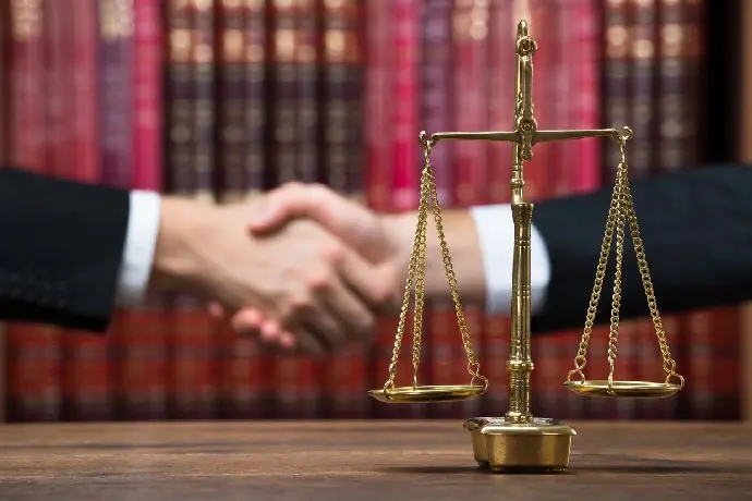 Two people shaking hands in the background with a balance scale in the foreground, set against a backdrop of bookshelves filled with legal books.