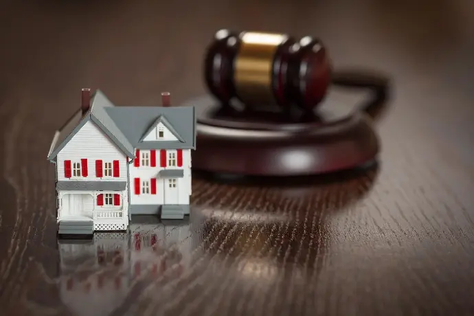 A miniature house sits next to a legal gavel on a wooden surface, symbolizing real estate law or property auction.