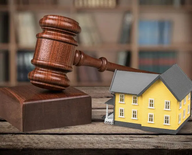 A judge's gavel next to a small model of a yellow house on a wooden surface, with bookshelves in the background.